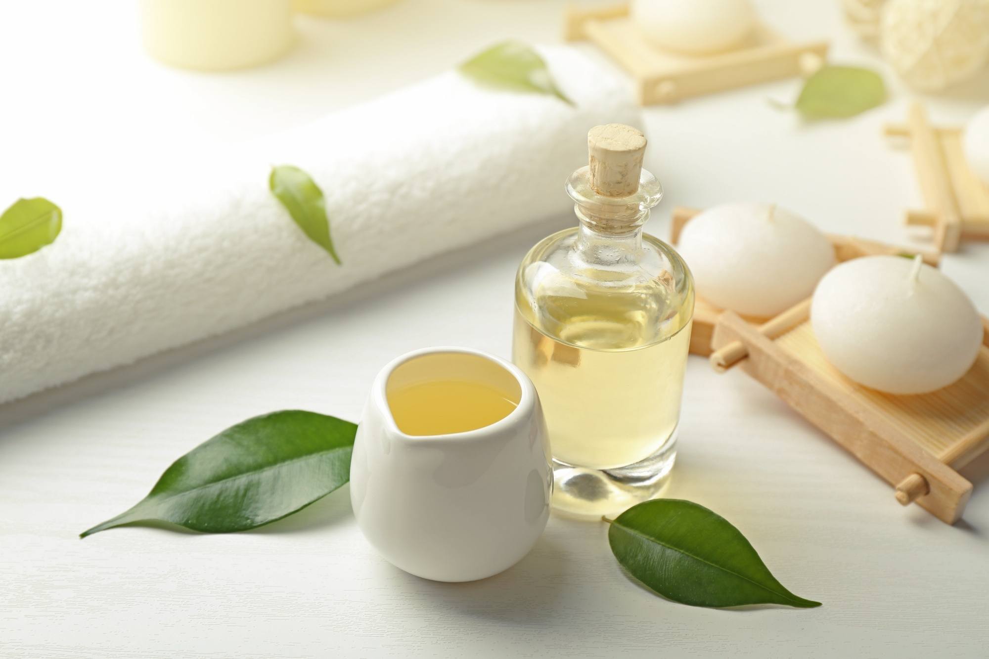 A bottle of tea tree oil on the table together with a small pitcher, white towel, and leaves