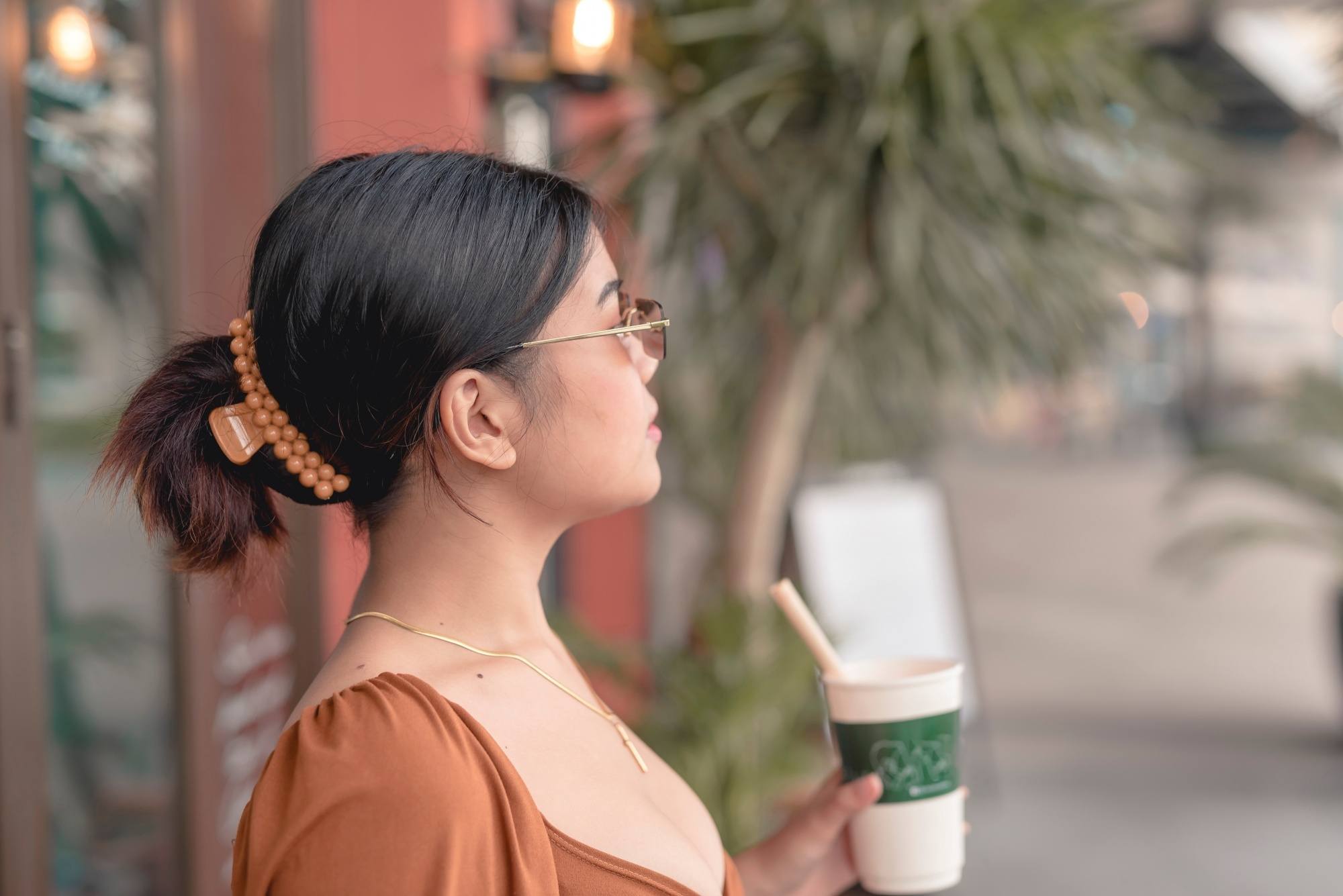 Asian woman with hair in a claw clamp standing outdoors and holding a cup of coffee.