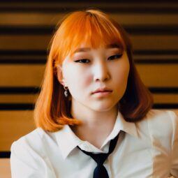 Asian woman with short ginger hair with bangs wearing a school uniform.